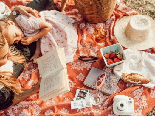 Мама с дочкой сидят на постеленном на землю покрывале и читают книгу.