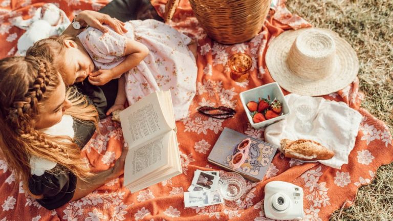 Мама с дочкой сидят на постеленном на землю покрывале и читают книгу.